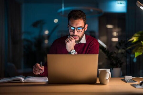 Mid adult Caucasian male concentrating on his work at a laptop in an office during nighttime with coffee and notebooks on his desk for online job