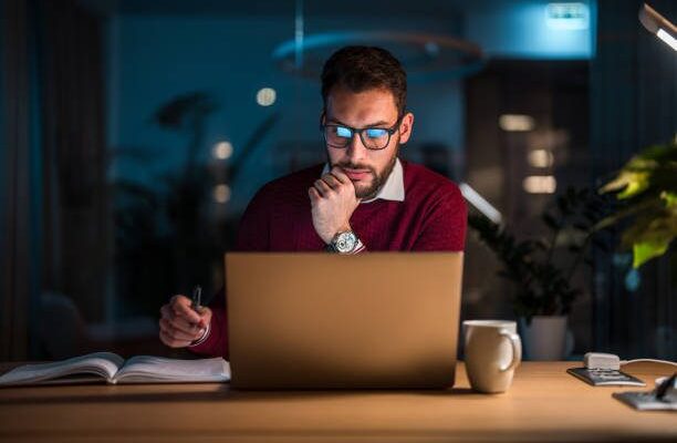 Mid adult Caucasian male concentrating on his work at a laptop in an office during nighttime with coffee and notebooks on his desk for online job